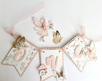 May include: A decorative banner with three pennants, featuring the letters "O", "N", and "E" in pink. The pennants are adorned with floral patterns, butterflies, and fairy-themed embellishments. A pink ribbon is attached for hanging.
