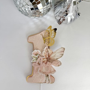 May include: A pink number one cake topper with a gold border, featuring a fairy illustration, a gold butterfly, and the name "Anya". The topper is designed for a first birthday celebration.