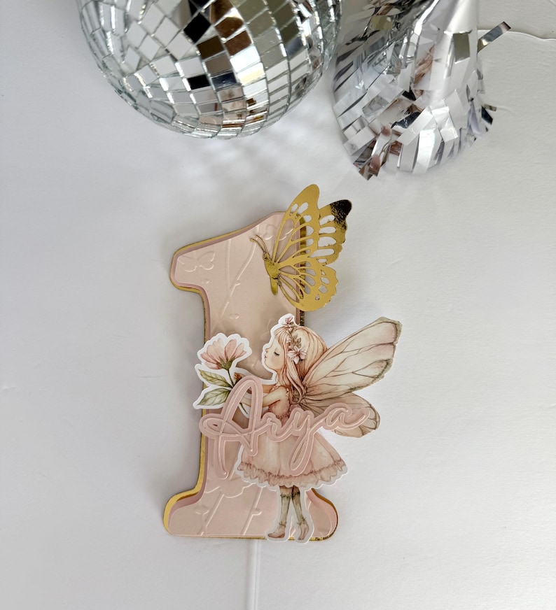 May include: A pink number one cake topper with a gold border, featuring a fairy illustration, a gold butterfly, and the name "Anya". The topper is designed for a first birthday celebration.