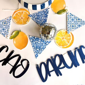 May include: A decorative banner featuring blue and white tile-patterned triangles, yellow lemon illustrations, and a central silver disco ball. The banner includes dark blue glittery script text that reads "CIAO DARLING" strung together on a white cord.