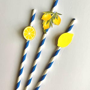 May include: Three paper straws with blue and white stripes, each adorned with a lemon-themed embellishment. One straw features a lemon slice, another has a cluster of lemons, and the third has a whole lemon.