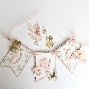 May include: A decorative banner with three pennants, featuring the letters "O", "N", and "E" in pink. The pennants are adorned with floral patterns, butterflies, and fairy-themed embellishments. A pink ribbon is attached for hanging.