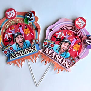 May include: Two custom cake toppers featuring the Danny Go! logo, a central portrait of a person wearing goggles, and a boombox graphic. Each topper displays the name AISLINN or MASON, the number 2, and decorative elements like flames and stars.