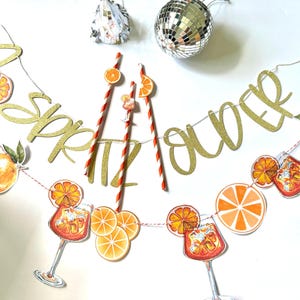 May include: A festive banner with gold glitter letters spelling "Spritz Over" is strung with orange slice and cocktail illustrations. Red and white striped straws and a disco ball add to the party theme.