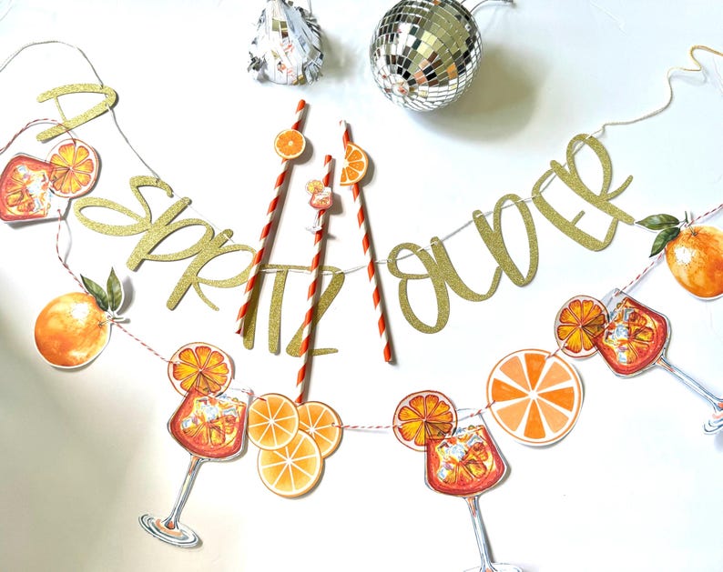 May include: A festive banner with gold glitter letters spelling "Spritz Over" is strung with orange slice and cocktail illustrations. Red and white striped straws and a disco ball add to the party theme.