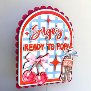 May include: A layered cake topper with a retro design. The topper features the text "Sage's READY TO POP!" in red and white, along with a cherry illustration and a soda bottle. The background has a blue and white checkered pattern.