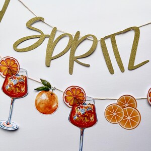 May include: A gold glitter banner spelling "Spritz" strung with a thin, natural-colored cord. The banner is decorated with illustrations of orange slices, oranges, and cocktails in stemmed glasses filled with ice and orange-colored liquid.