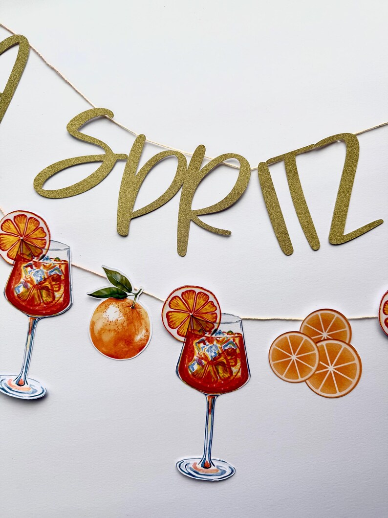 May include: A gold glitter banner spelling "Spritz" strung with a thin, natural-colored cord. The banner is decorated with illustrations of orange slices, oranges, and cocktails in stemmed glasses filled with ice and orange-colored liquid.