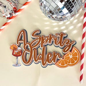 May include: A Spritz Older cake topper with a cocktail illustration and orange slice. The text is in brown, white, and blue, with a disco ball and red and white striped straw in the background.