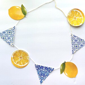 May include: A decorative garland featuring alternating paper cutouts of bright yellow lemons with green leaves, yellow lemon slices, and triangular pennants decorated with a blue and white intricate floral tile pattern, all strung together on a thin white cord.