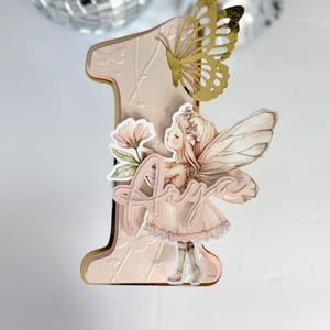 May include: A pink and gold number one cake topper with a butterfly and fairy design. The fairy holds a flower and the name "Aria" is written on the topper. The topper is made of layered paper and has a gold border.