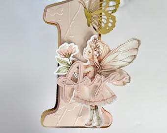 May include: A pink and gold number one cake topper with a butterfly and fairy design. The fairy holds a flower and the name "Aria" is written on the topper. The topper is made of layered paper and has a gold border.