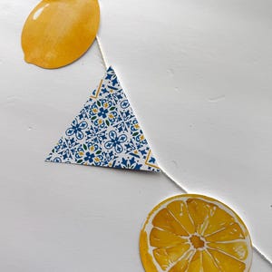 May include: A decorative garland featuring three distinct paper cutouts strung on a white cord: a whole yellow lemon with a green leaf, a triangular pennant with a blue and yellow floral tile pattern, and a circular slice of a yellow lemon.