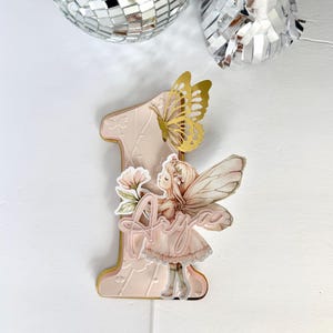 May include: A pink and gold number one cake topper with a fairy illustration and the name "Anya". The topper features a gold butterfly and floral accents. The background includes disco ball decorations.