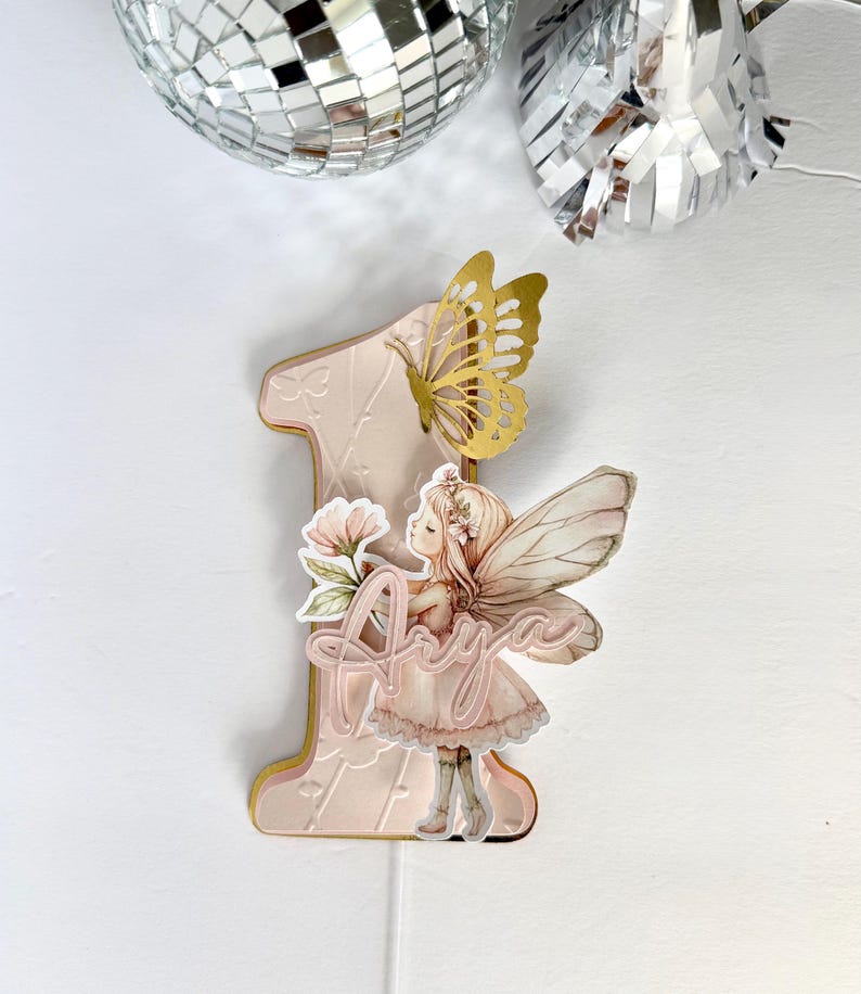May include: A pink and gold number one cake topper with a fairy illustration and the name "Anya". The topper features a gold butterfly and floral accents. The background includes disco ball decorations.