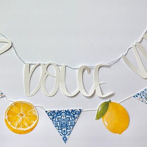 May include: A decorative banner featuring white cutout letters spelling LA DOLCE VITA, interspersed with yellow lemon illustrations and blue and white patterned triangular pennants, all strung together on a thin white cord for hanging display.