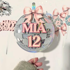 May include: A silver disco ball-shaped cake topper with the name "MIA" and the number "12" in pink. A pink bow is at the top. Additional pink bows and disco ball decorations are in the background. The words "Happy Birthday" are also visible.