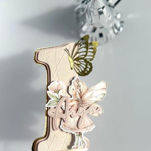 May include: A decorative number one cake topper with a fairy and butterfly design. The number is light pink with gold trim, featuring a fairy illustration and the name "Aiya". A gold butterfly is attached to the top of the number. A silver foil decoration is in the background.