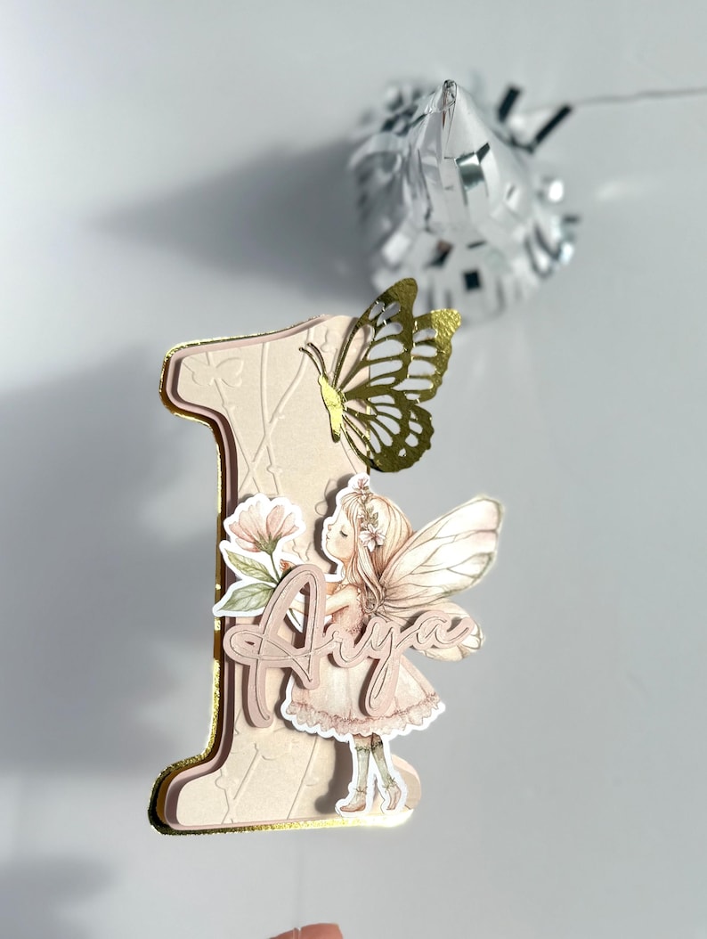 May include: A decorative number one cake topper with a fairy and butterfly design. The number is light pink with gold trim, featuring a fairy illustration and the name "Aiya". A gold butterfly is attached to the top of the number. A silver foil decoration is in the background.