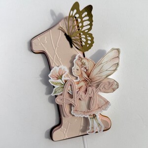 May include: A pink number one cake topper with a butterfly and fairy design. The butterfly is gold with intricate cutouts. The fairy is holding flowers and the name "Amy" is written on the topper. The topper is designed for a first birthday.