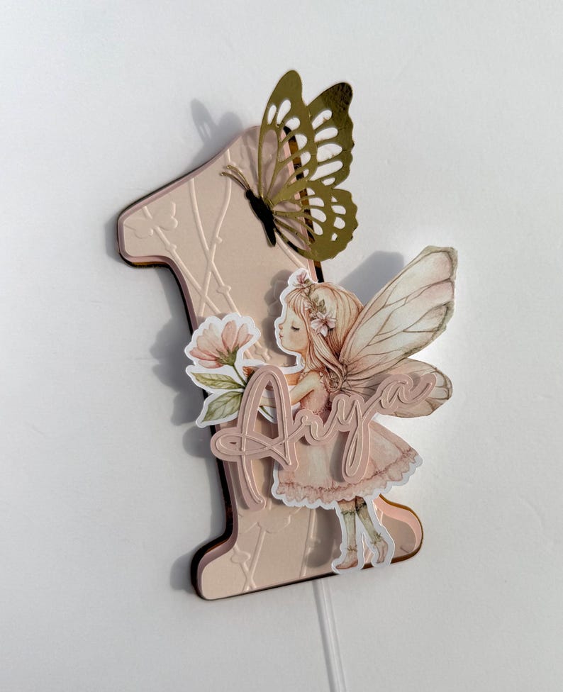 May include: A pink number one cake topper with a butterfly and fairy design. The butterfly is gold with intricate cutouts. The fairy is holding flowers and the name "Amy" is written on the topper. The topper is designed for a first birthday.