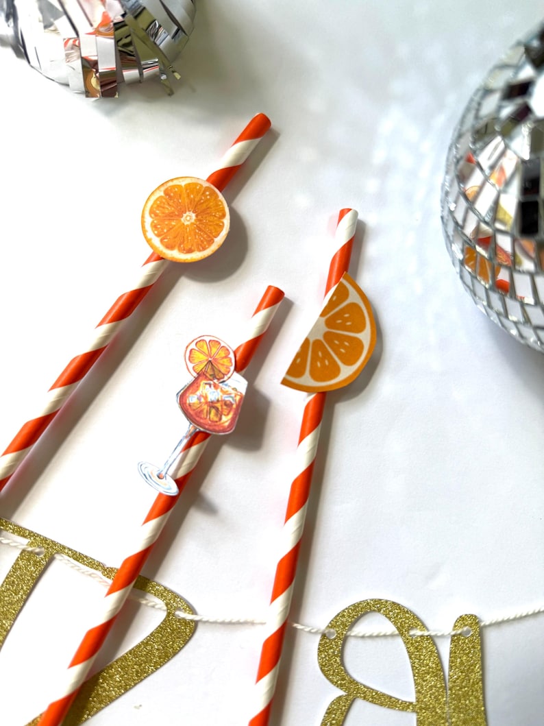 May include: Close-up of orange and white striped paper straws with orange slice decorations. The straws are arranged on a white surface with a disco ball and a gold glitter banner in the background. Party supplies.