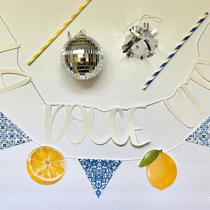 May include: A decorative banner featuring the text LA DOLCE VITA in white script, interspersed with yellow lemon illustrations, green leaves, and blue and white patterned triangular pennants, accompanied by a disco ball and two striped straws.