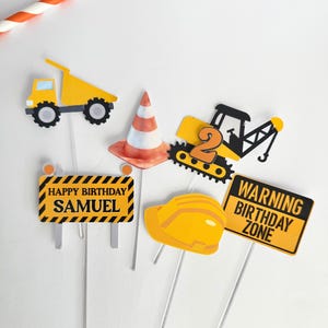 May include: A collection of construction-themed cake toppers. Includes a yellow dump truck, traffic cone, excavator with the number 2, a hard hat, and signs that read "Happy Birthday Samuel" and "Warning Birthday Zone".