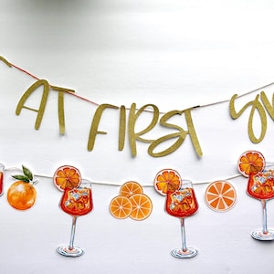 May include: A banner with the words "LOVE AT FIRST SPRITZ" in gold glitter lettering. Below the banner are illustrations of Aperol Spritz cocktails, orange slices, and whole oranges, all on a white background.