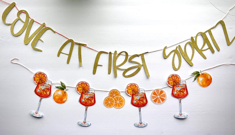 May include: A banner with the words "LOVE AT FIRST SPRITZ" in gold glitter lettering. Below the banner are illustrations of Aperol Spritz cocktails, orange slices, and whole oranges, all on a white background.