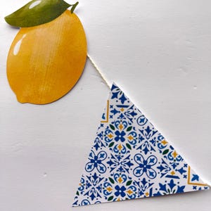 May include: A decorative garland featuring a yellow lemon cutout with a green leaf and a triangular pennant with a blue, yellow, and green geometric tile pattern, both strung together on a thin white cord against a plain white background.