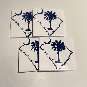 May include: Four white ceramic coasters with a blue outline of the state of South Carolina and a palmetto tree. The text "South Carolina" is written on each coaster.