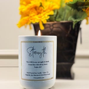 Puede incluir: Una vela blanca encendida con la palabra "Strength" y un versículo bíblico impreso en la etiqueta. La vela está frente a un recipiente de metal marrón oscuro lleno de flores amarillas. La vela mide 10 cm de alto.