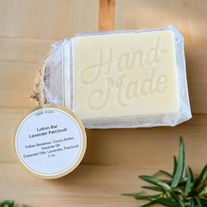 Op de afbeelding: Een rechthoekige, crèmekleurige lotionbar in een gaaszak, met de tekst "Hand-Made". Een rond etiket leest "Lotion Bar Lavender Patchouli" met ingrediënten. De bar is 56.7 gram en bevat gele bijenwas, cacaoboter, sesamolie en essentiële oliën.