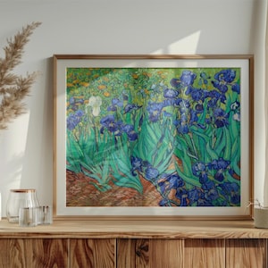 Puede incluir: Impresión enmarcada de una pintura que representa lirios azules y hojas verdes. La obra de arte está en un marco de madera y se exhibe sobre un mueble de madera. Un jarrón blanco con plantas secas y otros objetos decorativos están en el mueble.