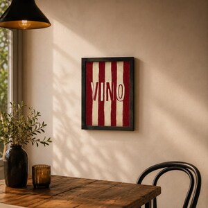 Op de afbeelding: Een ingelijst kunstwerk met een rood-witte gestreepte achtergrond en het woord "VINO" in witte letters. Het kunstwerk wordt tentoongesteld aan een beige muur, boven een houten tafel. Een zwarte vaas en een glazen kandelaar staan op de tafel.