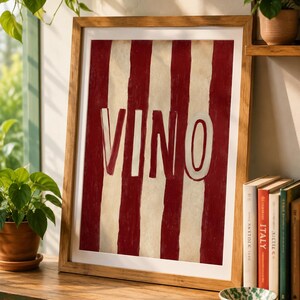 Op de afbeelding: Een ingelijst kunstwerk met een rood-witte gestreepte achtergrond en het woord "VINO" in wit. De houten lijst complementeert het kunstwerk, dat op een houten oppervlak wordt getoond. Een decoratief stuk voor een huis.