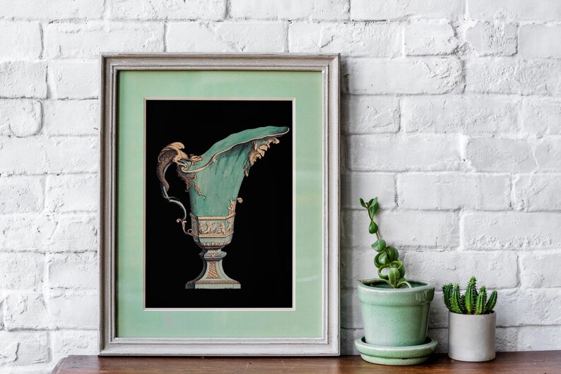 May include: Framed print of an antique teal and gold decorative urn against a black background. The urn has ornate details and a curved spout. The frame is a light green color. The print is displayed on a wooden surface with a white brick wall backdrop.