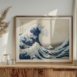 May include: A framed print of Hokusai's "The Great Wave off Kanagawa". The artwork shows a large wave with a boat and Mount Fuji in the background. The print is in a wooden frame and displayed on a wooden cabinet.