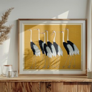 May include: Framed artwork featuring six Japanese cranes in a row, set against a golden yellow background. The birds have white necks, black and gray wings, and long, thin legs. The artwork is displayed on a wooden cabinet.