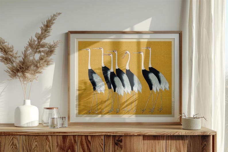 May include: Framed artwork featuring six Japanese cranes in a row, set against a golden yellow background. The birds have white necks, black and gray wings, and long, thin legs. The artwork is displayed on a wooden cabinet.