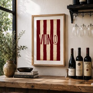 Op de afbeelding: Een ingelijste print met rode en cr&egrave;mekleurige strepen en het woord "VINO" in grote letters. De print wordt getoond op een houten bar met wijnflessen en glazen, wat wijst op een wijn-thema.