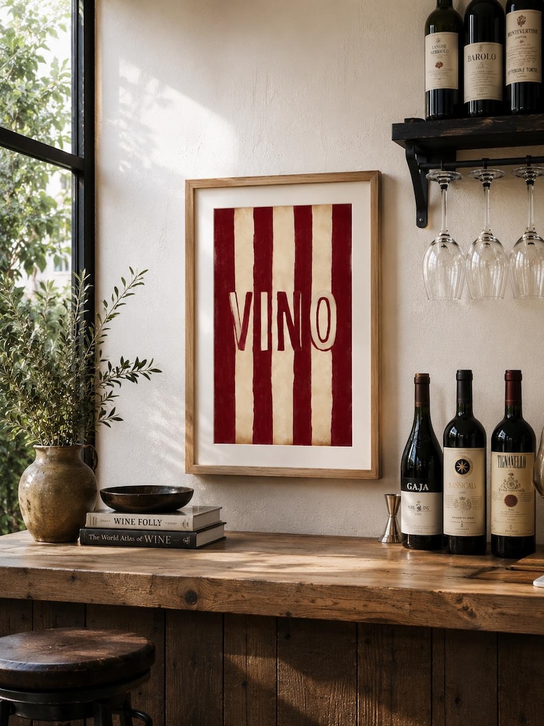 Op de afbeelding: Een ingelijste print met rode en cr&egrave;mekleurige strepen en het woord "VINO" in grote letters. De print wordt getoond op een houten bar met wijnflessen en glazen, wat wijst op een wijn-thema.