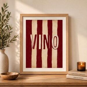 Op de afbeelding: Houten ingelijst kunstwerk met een rood en cr&egrave;me gestreept ontwerp. Het woord "VINO" wordt weergegeven in een vetgedrukte, witte, omlijnde letter. Het kunstwerk wordt getoond op een houten oppervlak.
