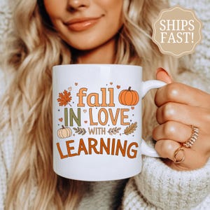 May include: A white ceramic mug featuring the orange and green text "fall IN LOVE WITH LEARNING" surrounded by illustrations of orange pumpkins, a maple leaf, and small hearts. The mug has a curved handle and a smooth, glossy finish.