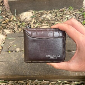 Op de afbeelding: Een bruine leren portemonnee met ritssluiting. De portemonnee heeft een textuur, krokodilachtig patroon op het bovenste gedeelte en de tekst "CARRKEN STYLE LEATHER" is zichtbaar.