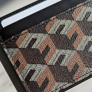 Może przedstawiać: Zbliżenie na etui na karty z geometrycznym wzorem w odcieniach brązu, beżu i szarości. Etui na karty ma czarną skórzaną obwódkę, a biała karta jest częściowo widoczna. Wzór jest przeszyty kontrastującą nicią.