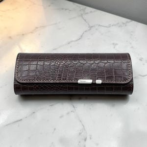 May include: A rectangular dark brown case with a textured crocodile skin pattern. The case features a flap closure secured by a slim, horizontal silver-toned metal clasp centered on the front edge, with visible stitching along the perimeter.