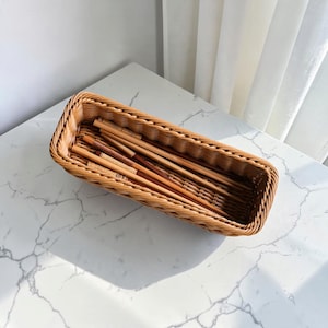 May include: A rectangular woven wicker basket with a light brown finish, featuring a braided rim and a lattice-style base. Inside the basket, several wooden chopsticks are arranged horizontally, showcasing varying shades of natural wood grain.