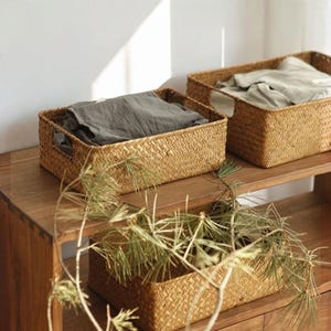May include: Three rectangular woven seagrass baskets with integrated side handles. Two baskets are placed on a wooden shelf, each containing folded fabric, while a third empty basket sits below, partially obscured by pine branches.